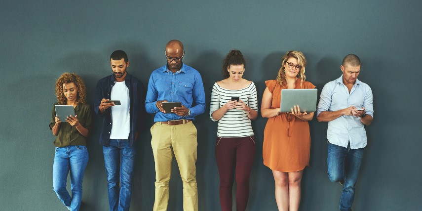 A row of six people stand against a wall, all looking at their devices.