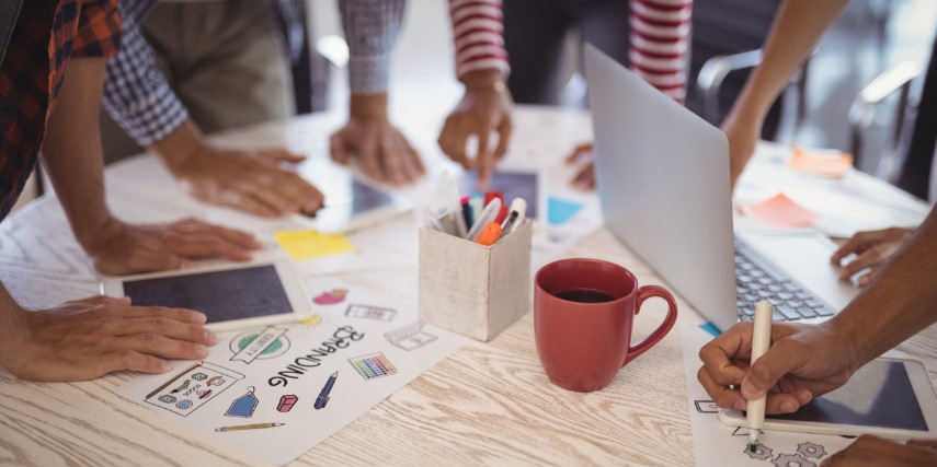 Table surface littered with a coffee mug, pen cup and papers with graphics as people gather around it, working. 