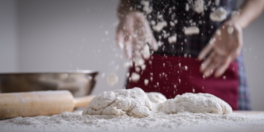A pile of floury dough is on a table surface while a baker stands behind it, tossing in more flour. A bowl and rolling pin are on the table behind the dough.