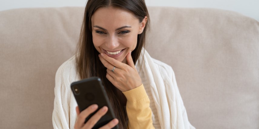 A woman with long, dark hair sits on a couch, smiling at something she is watching on a cell phone.
