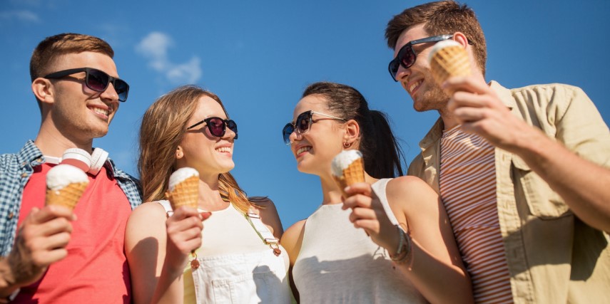 Four people wearing sunglasses stand close together eating ice cream and smiling.