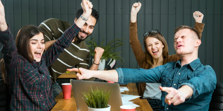 Four people sit around a table, cheering about something they see on a laptop.