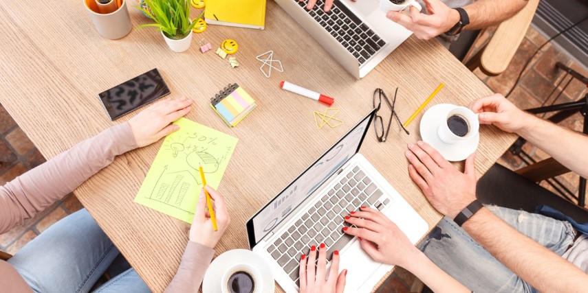 Overhead view of a conference table with two laptops, tablets of paper and people's hands as they collaborate.