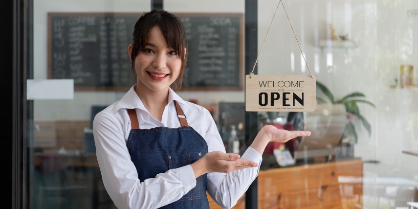 A woman in a white shirt and apron shows a sign that says, "Welcome: Open" in front of a coffee shop.