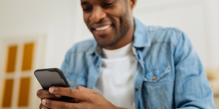 A man in a denim shirt is looking at a smartphone and smiling.