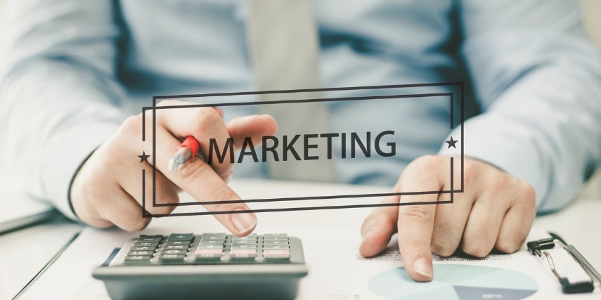 A person is sitting at a desk using a calculator and the word "marketing" is in the foreground of the image.