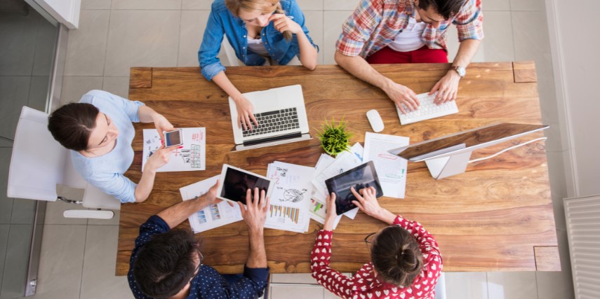 A group of people are gathered around a table with charts, laptops and calculators, discussing marketing budgets.