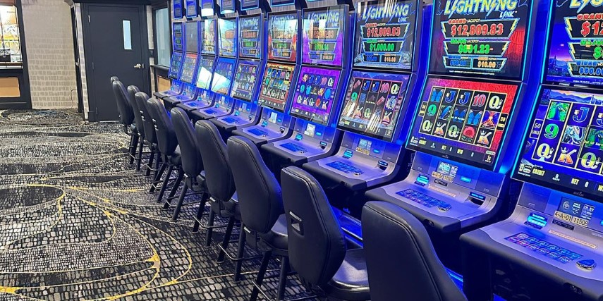 A row of primarily blue slot machines with a chair in front of each machine in a carpeted room.