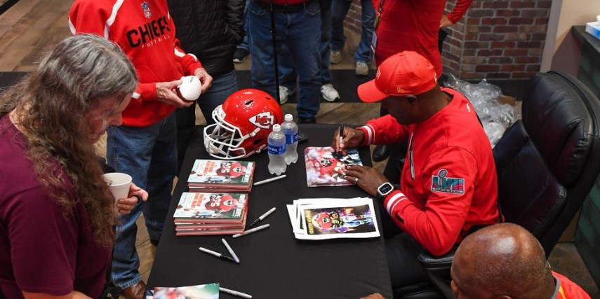 People line up to receive autographs at a table. A Kansas City Chiefs helmet and other team items are displayed on the table.