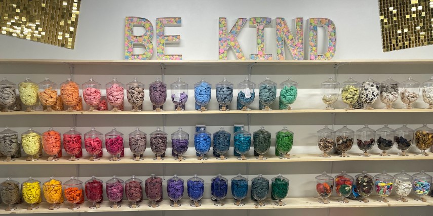 Rows of lidded glass jars display various balloon colors. A sign above the selection says, "Be Kind."