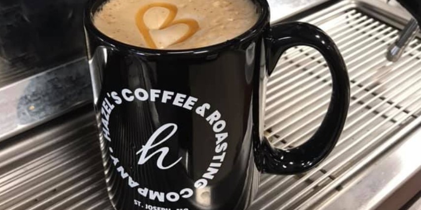 A black coffee mug with white writing on the side circling a white italic "H." The surface of the coffee is foamy.