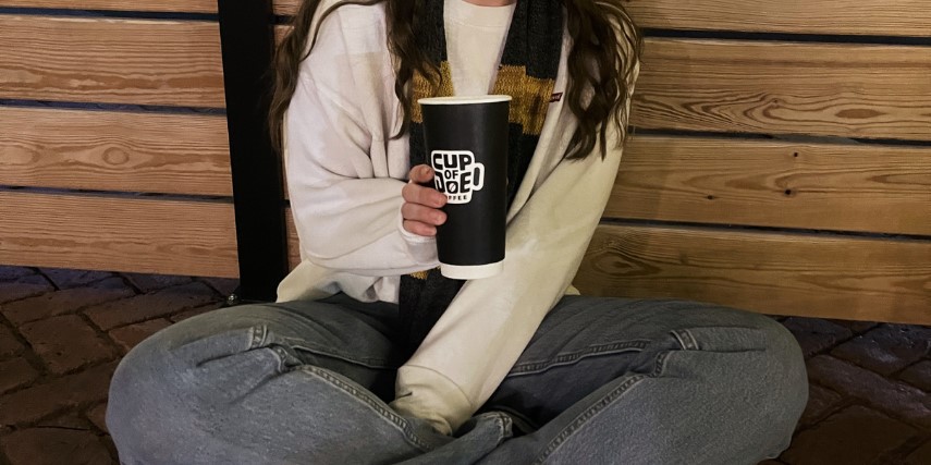 A woman sits cross-legged on the floor, holding out a brown paper coffee cup that says "Cup of Joe." 