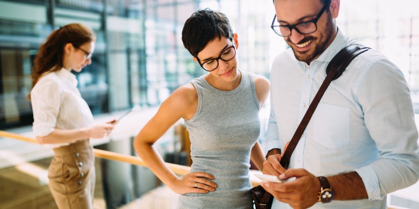 A man stands in an office looking at a smartphone while a woman stands next to him looking at the same phone.