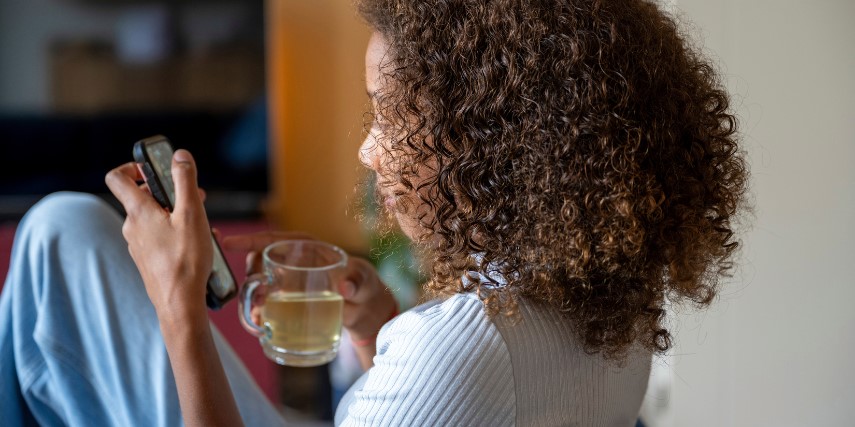 A young woman in a white shirt and blue pants sits holding a cup of tea in one hand and a smartphone in the other.