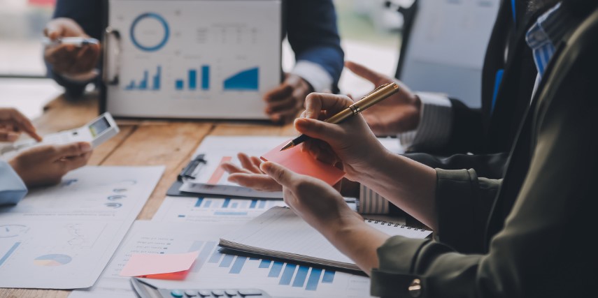 At a meeting table, a person at the head of the table holds up a graph to show the team.