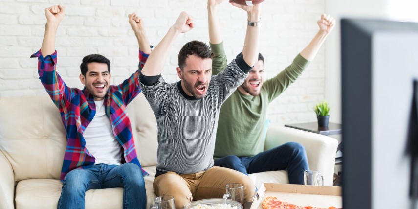Three men sit on a couch with their arms raised in a cheering position.