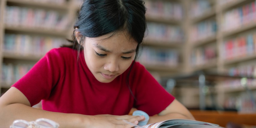 A young girl in a red shirt sits at a table looking at a book with bookshelves in the background.