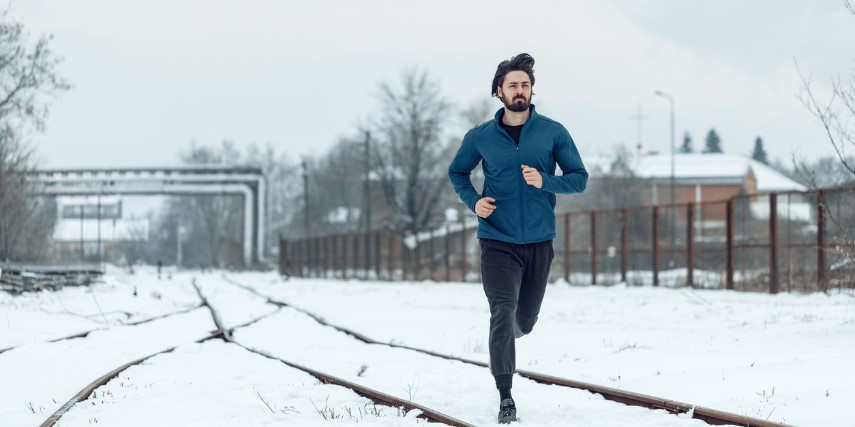 A young man dressed in dark fitness clothing runs along snow-covered train tracks.