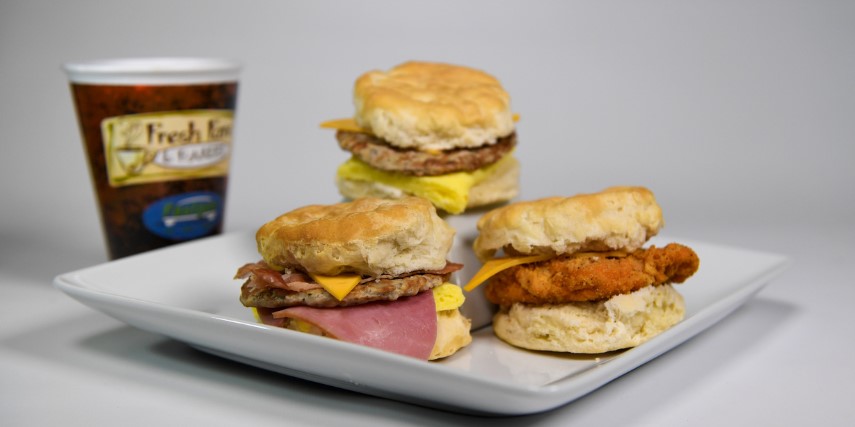 Three biscuit breakfast sandwiches are arranged on a white plate next to a cup of coffee.