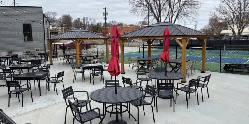 A patio with umbrella tables and chairs with pickleball courts in the background.