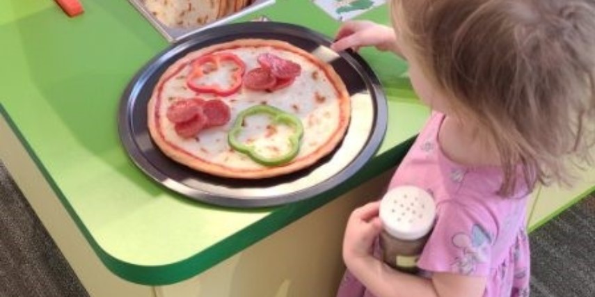 A young girl makes a pretend pizza at a green counter.