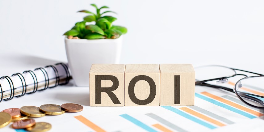 The letters "ROI" in black ink on wood blocks sit on a desk by a notebook and a potted plant.
