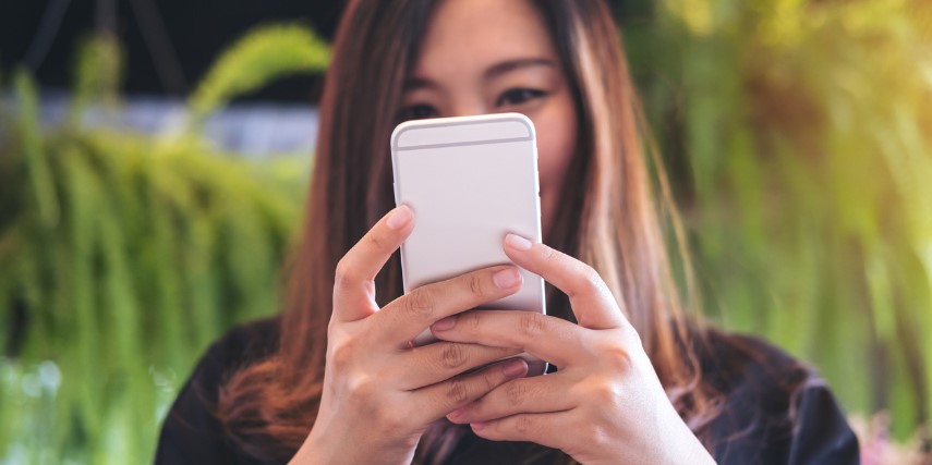 A woman in the background holds her smartphone in the foreground with both hands.