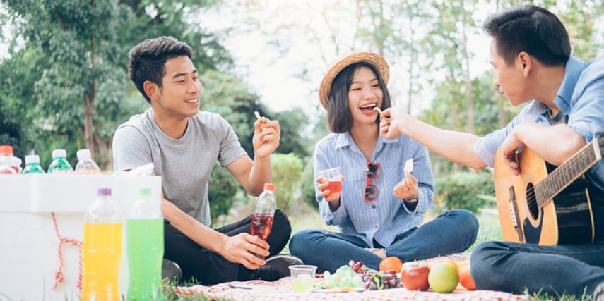 Three people sit on a blanket enjoying food and drinks while one person plays guitar.