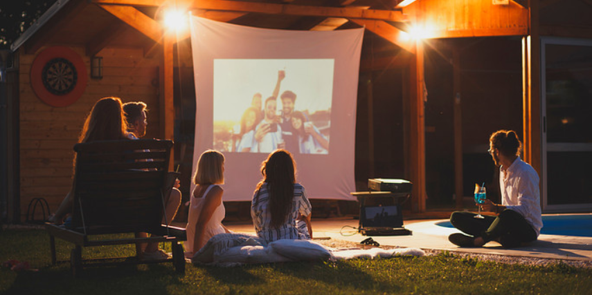 Friends sit watching a makeshift outdoor movie projected on a sheet. 