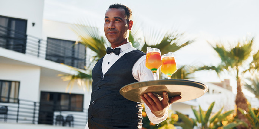 A waiter is outside, carrying two orange and yellow cocktails on a tray.