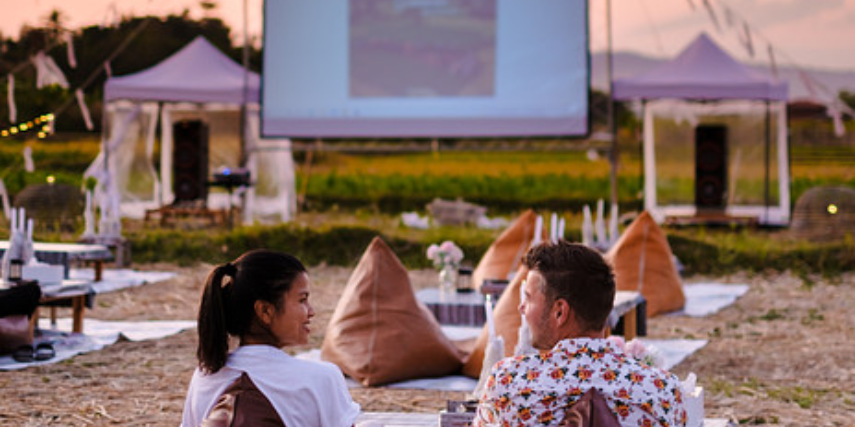 A couple sits on a beach watching an outdoor movie screen.