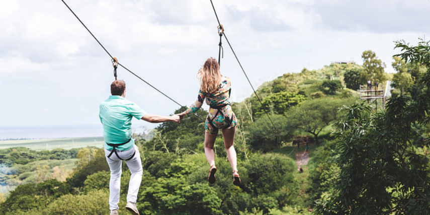 A couple ziplining through trees while holding hands.